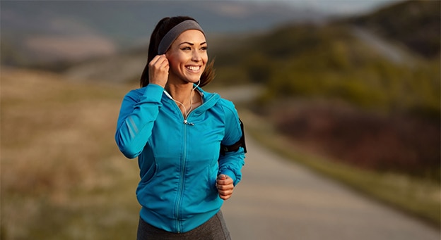 Smiling woman running on a rural road while wearing a blue jacket, headband, and earphones.