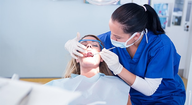Female dentist wearing blue scrubs and protective gear examining a female patient's teeth in a clinic