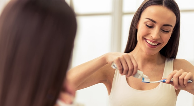 Smiling woman applying toothpaste to a toothbrush in front of a bathroom mirror