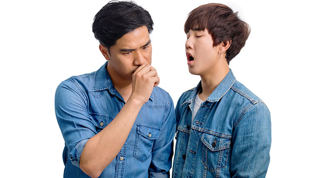 Man checking his breath and another man reacting.
